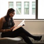 A thoughtful woman reading a book while sitting by a window indoors.