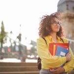 A young woman with curly hair and a yellow shirt stands confidently outside a historical building, holding books.