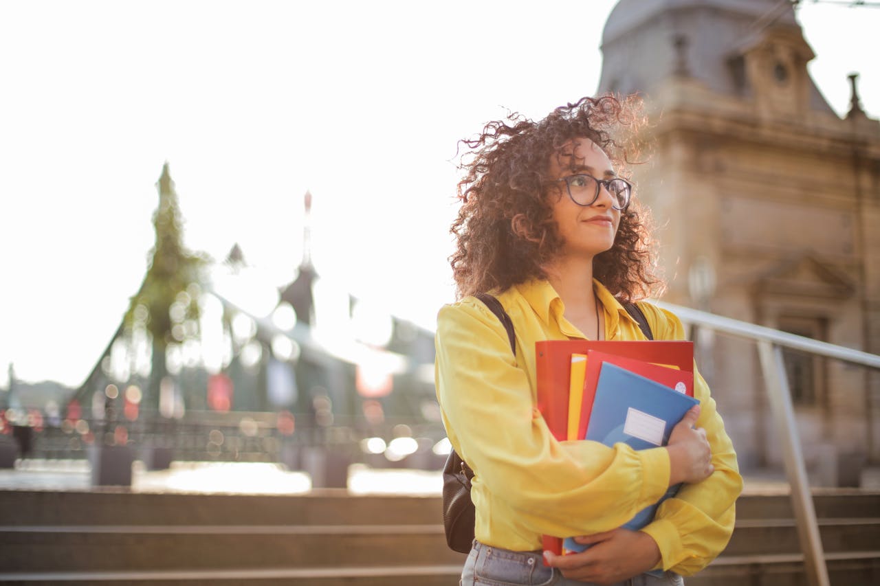 A young woman with curly hair and a yellow shirt stands confidently outside a historical building, holding books.